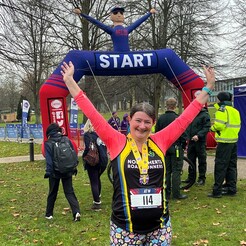 Clare wearing a running race medal with hands in the air in a V in front of the inflatable startfinish gantry that also has an inflatable man on top in the same pose